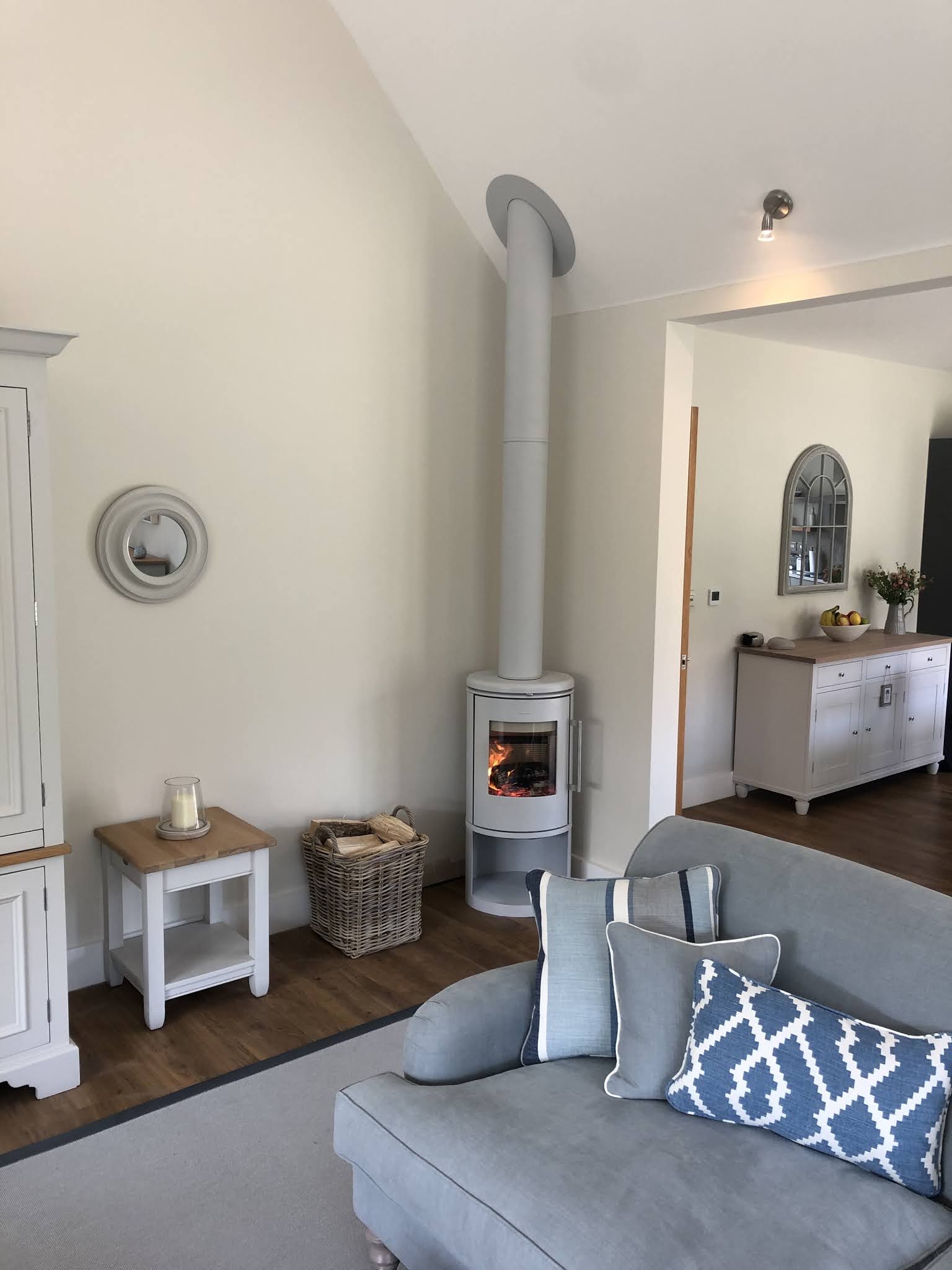A pale grey round wood burner with a tall freestanding flue pipe in a bright vaulted living room